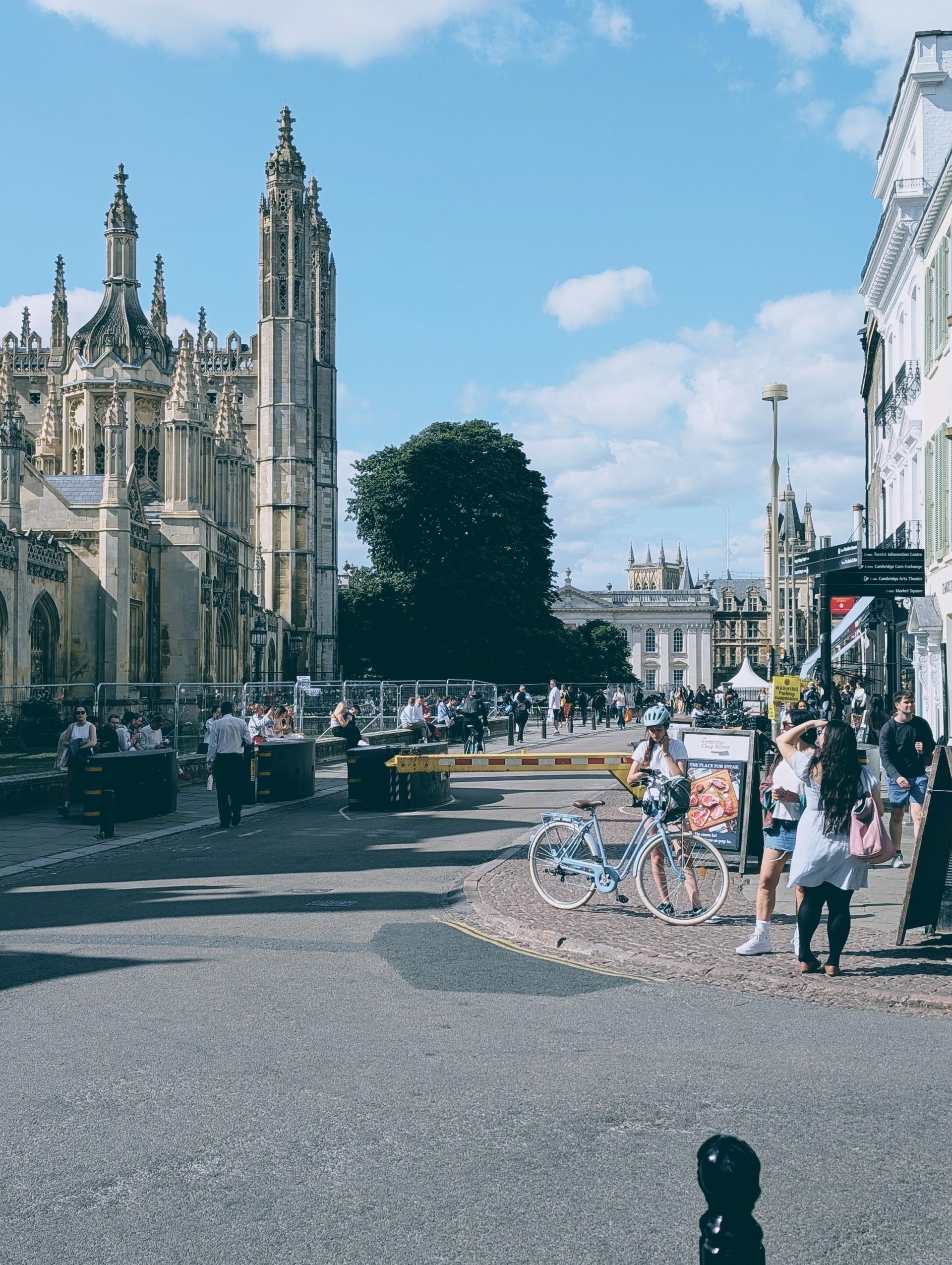 A busy street scene near King's College in Cambridge, with pedestrians, cyclists and Gothic spires of a UK university under a bright sky.