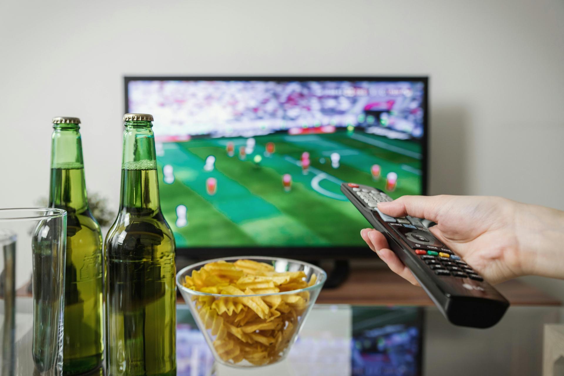A viewer holds a television remote while a football match plays on screen beside snacks and drinks