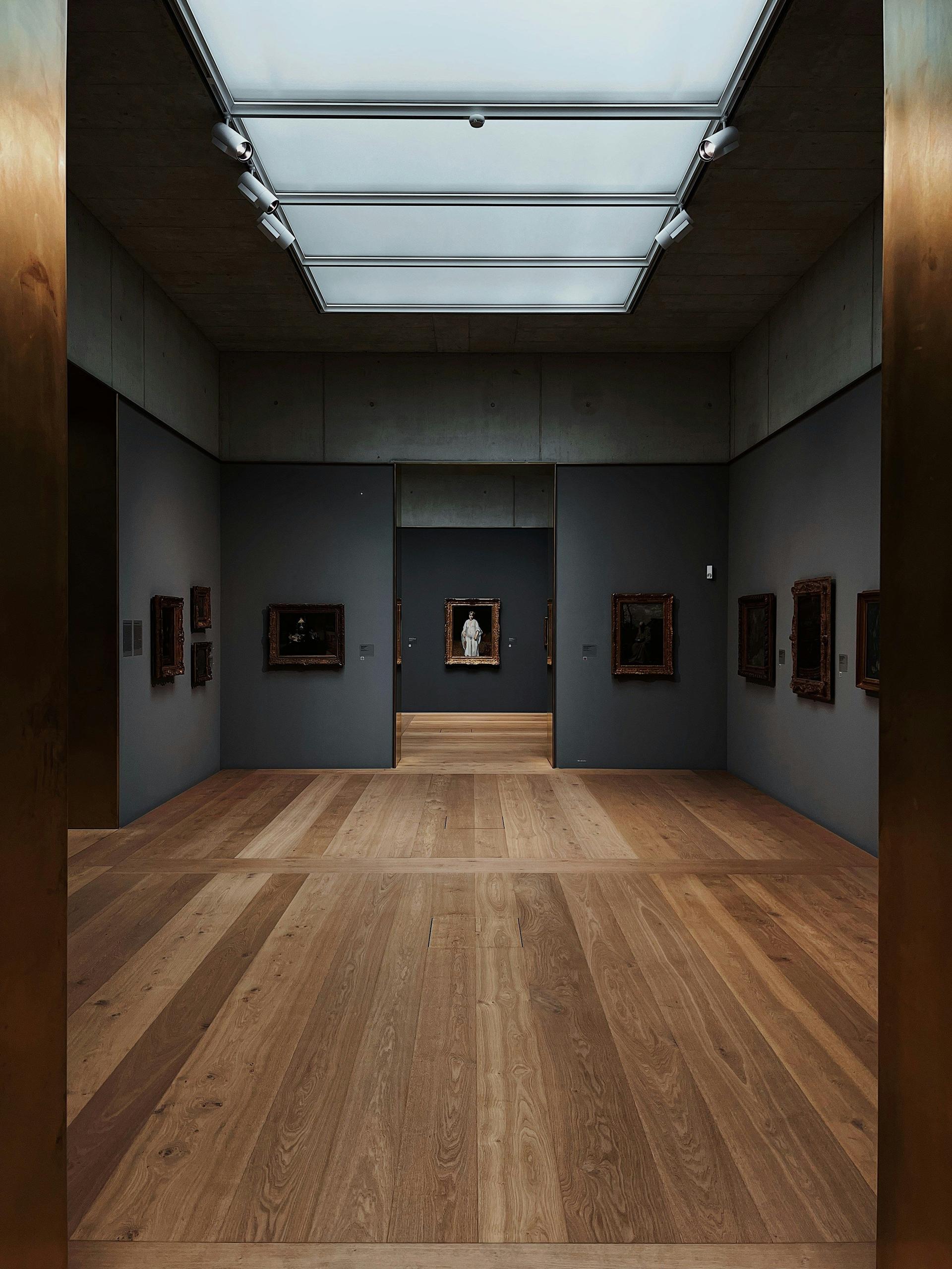 Visitors viewing framed paintings on white walls inside a contemporary art gallery with wood flooring