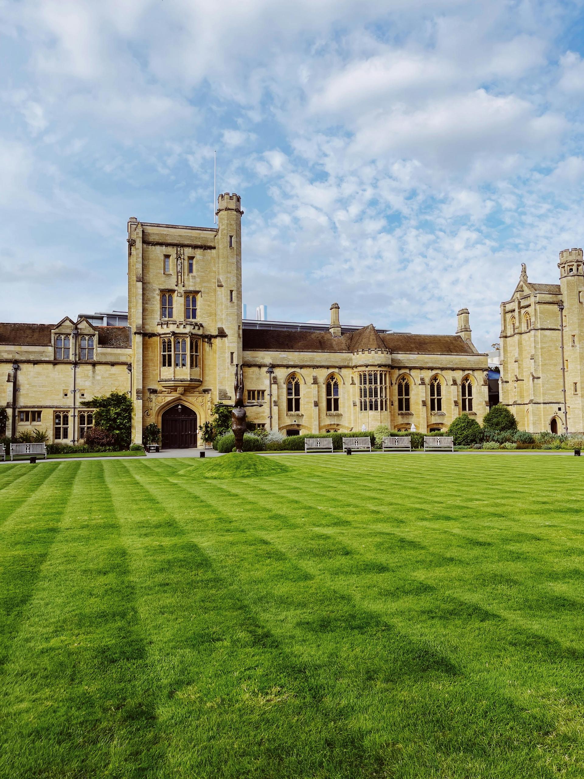 Historic British university building with a wide green lawn under a blue sky, evoking UK higher education.
