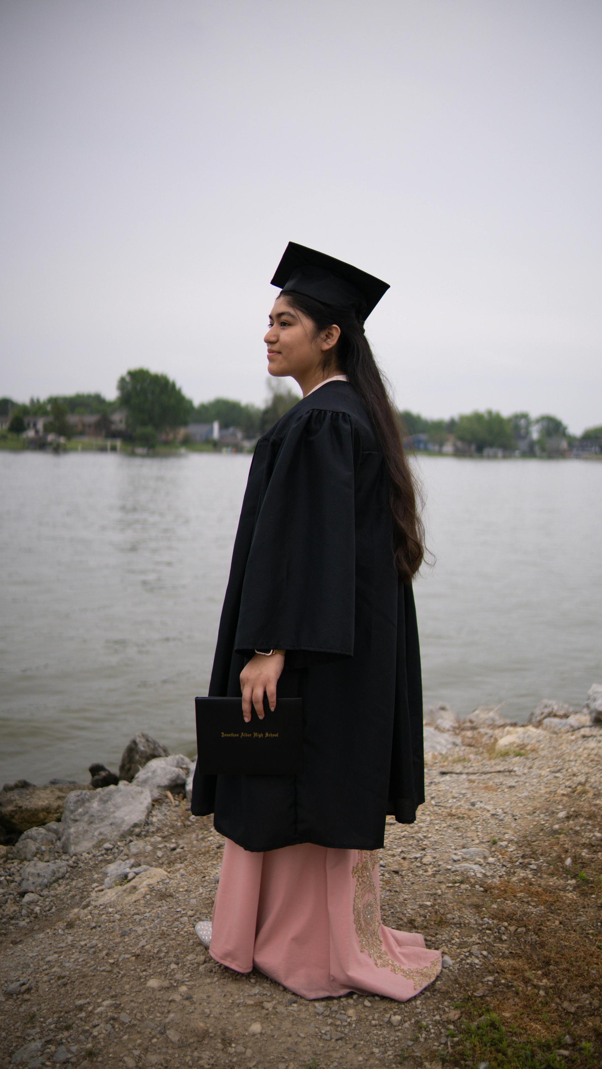 A young graduate in cap and gown stands by a quiet lakeside, evoking the threshold between student life in America and what comes next.