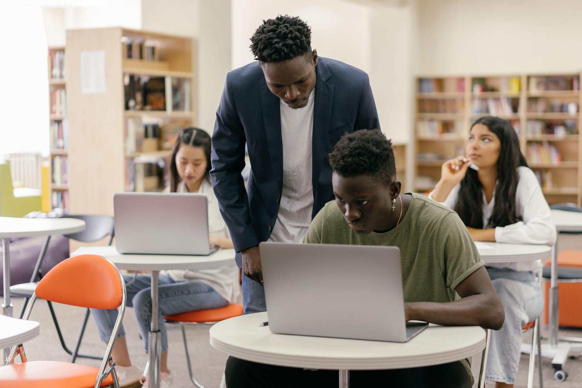 A teacher leans over students working on laptops in a classroom, guiding their learning