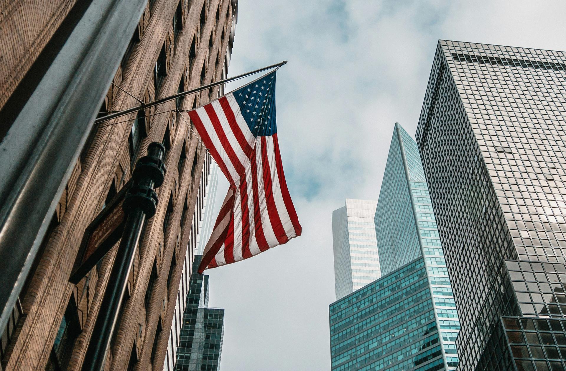 An American flag hangs from a building among city skyscrapers, symbolising citizenship and belonging in the United States.