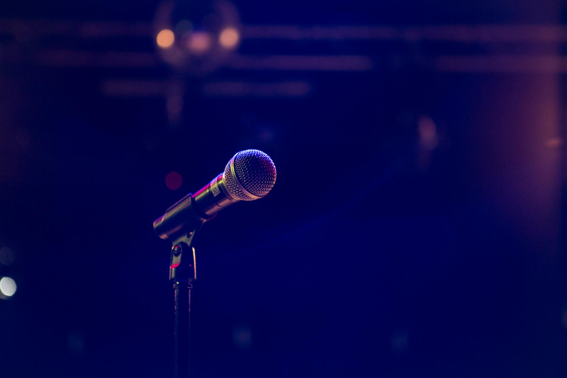 A microphone on a stand under stage lighting against a dark background, evoking a stand-up comedy stage