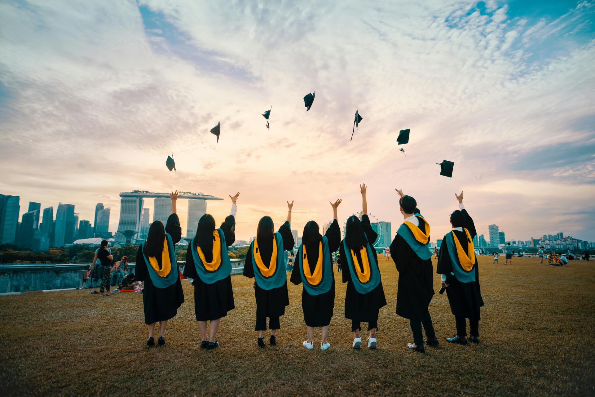 Graduating students in caps and gowns throw their mortarboards into the air against a city skyline at sunset