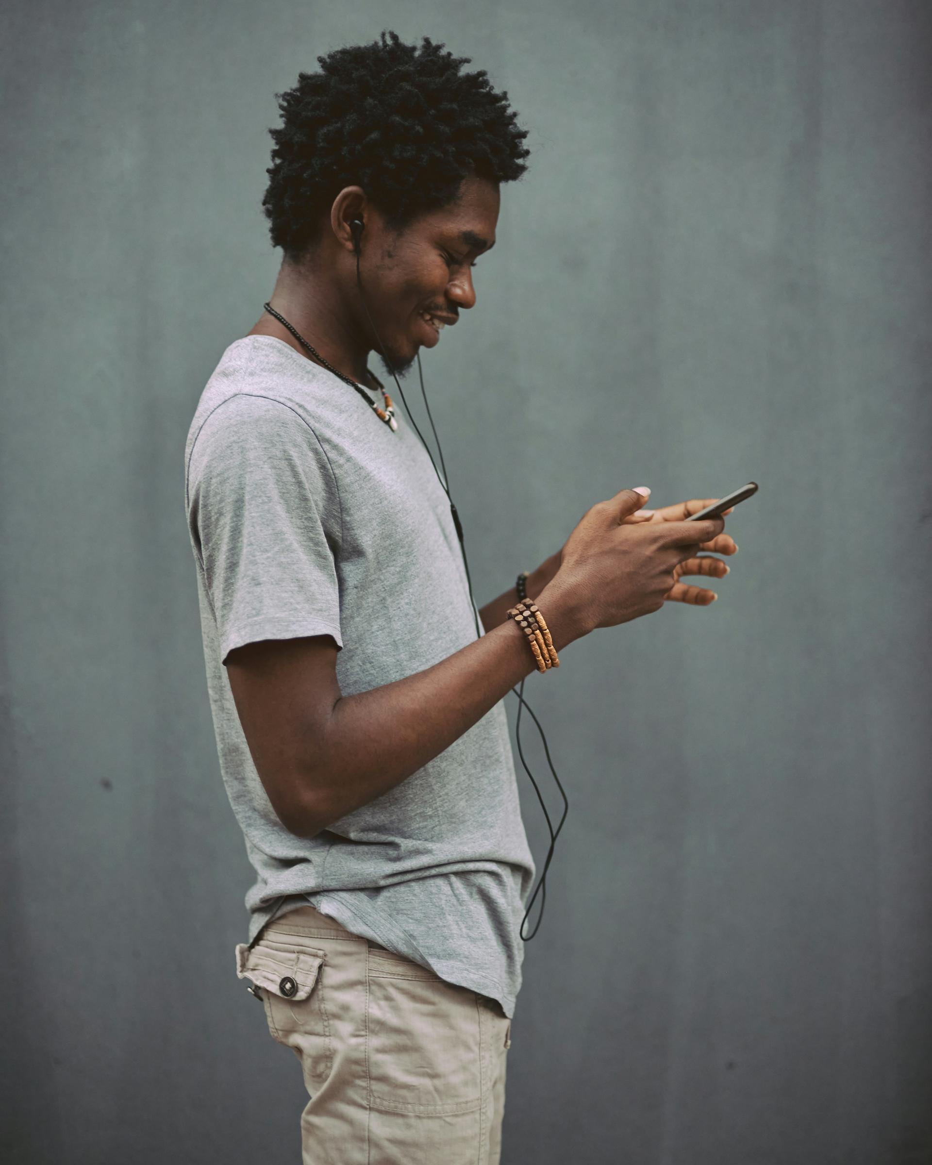 A young African man in a grey t-shirt checks his smartphone screen with earbuds in, standing against a concrete wall.