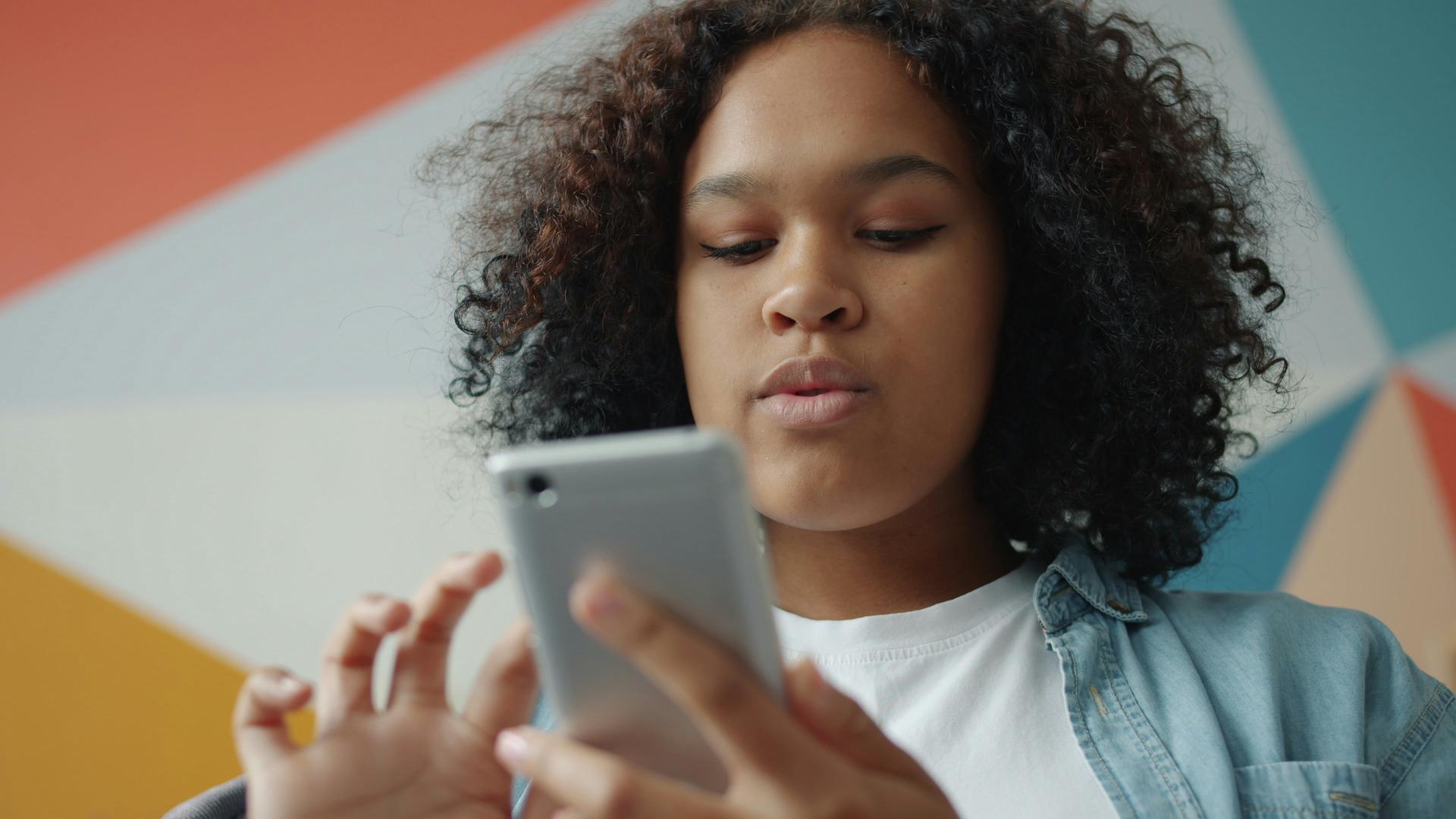 A young person looks at a smartphone screen lit against a colourful background, illustrating teenage social media use.