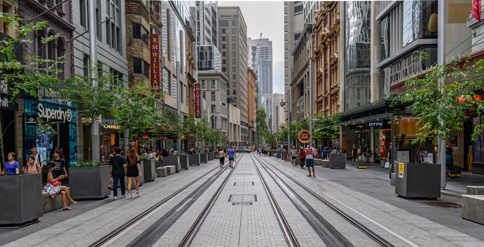 A street-level view of George Street in Sydney's central business district, with pedestrians, light-rail tracks and tall office towers under daylight.