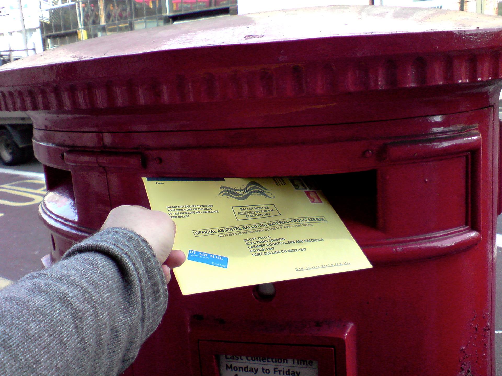 A single ballot paper being dropped into a sealed transparent ballot box at a polling station.