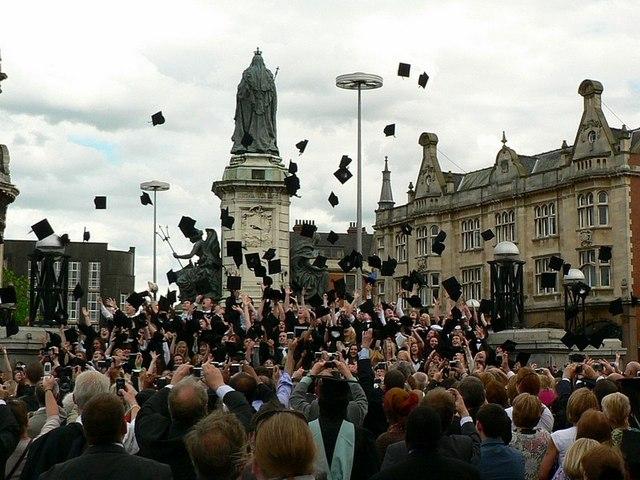 Graduating UK university students throw their black mortarboard caps into the air at an outdoor ceremony.