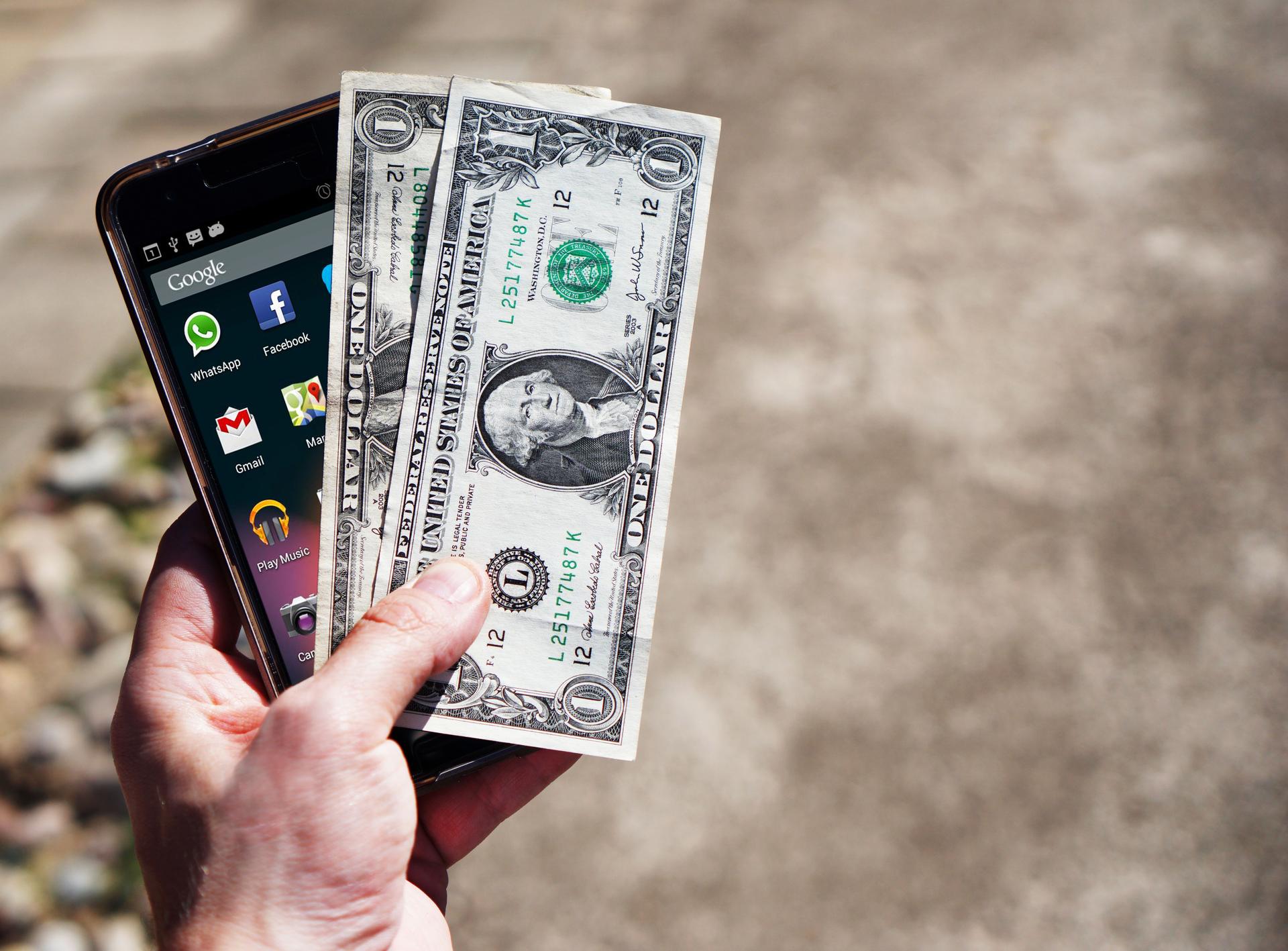 A hand holds a smartphone and US dollar banknotes, illustrating mobile-money and cross-border remittances.