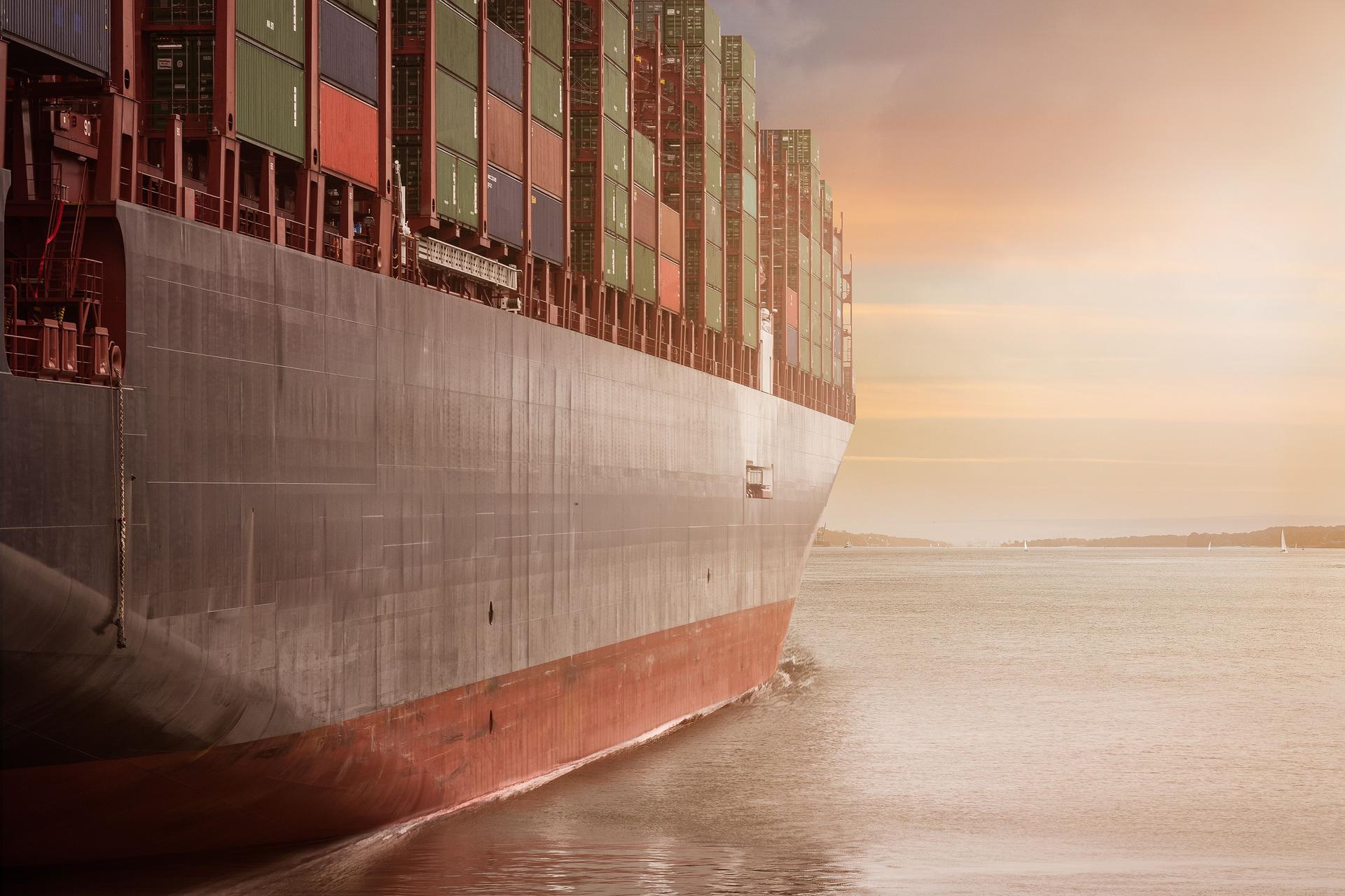A large cargo ship loaded with containers sails through calm ocean waters at sunset.