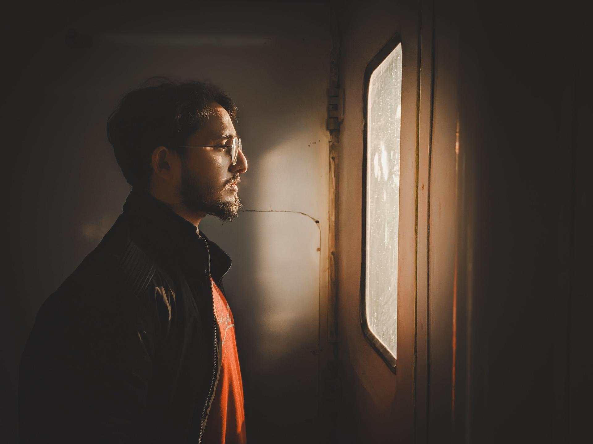 A man stands indoors in warm light, looking thoughtfully out of a window, conveying quiet reflection.