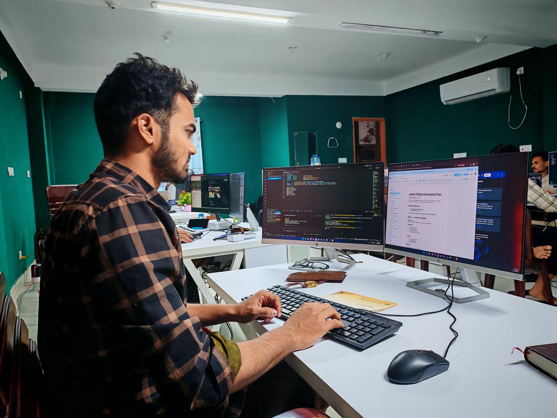A software developer works on code at a dual-monitor desk in a modern office setting.