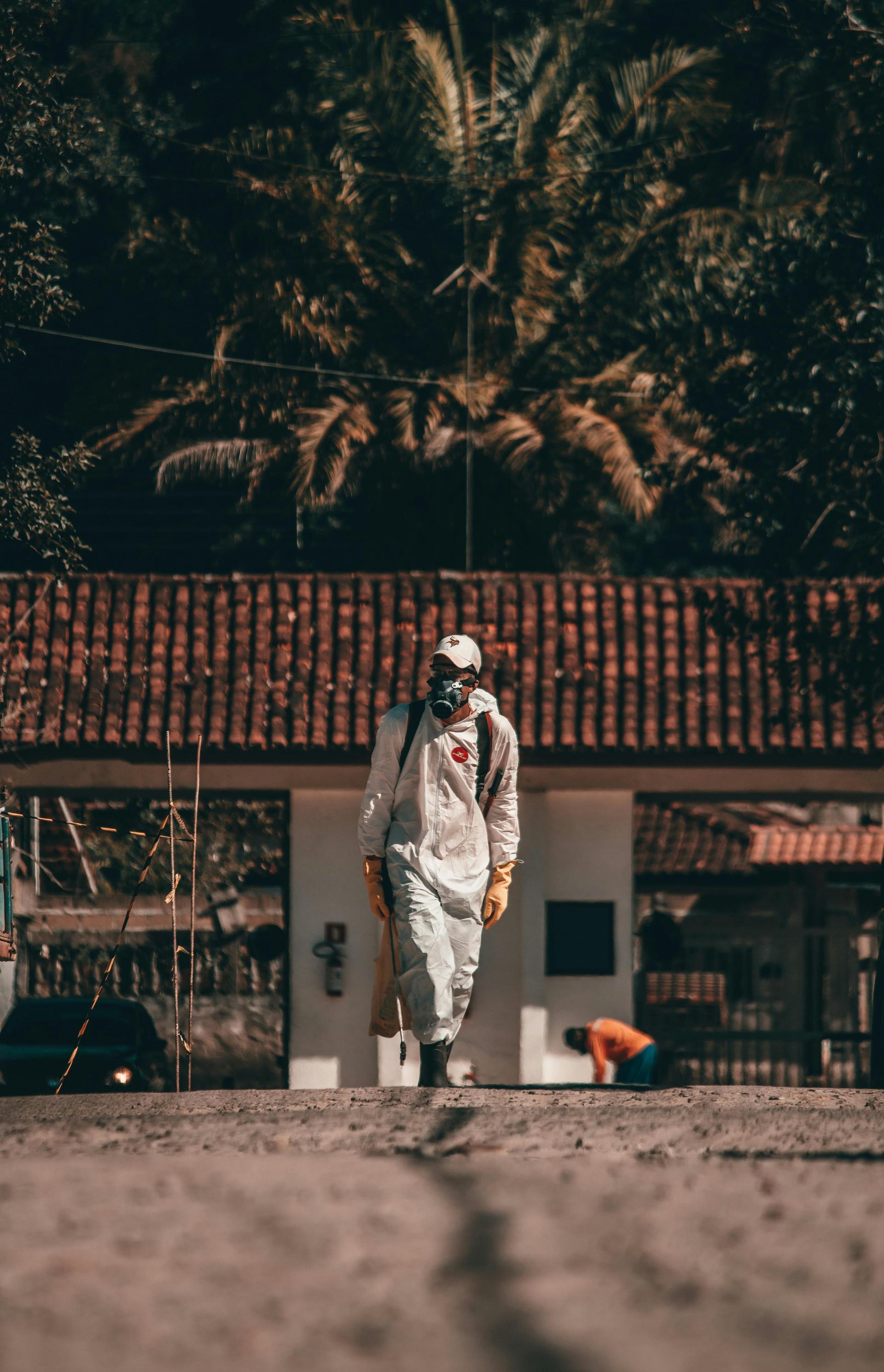 A worker in a full hazardous-materials suit and respirator walks through a tropical rural setting during an epidemic response.