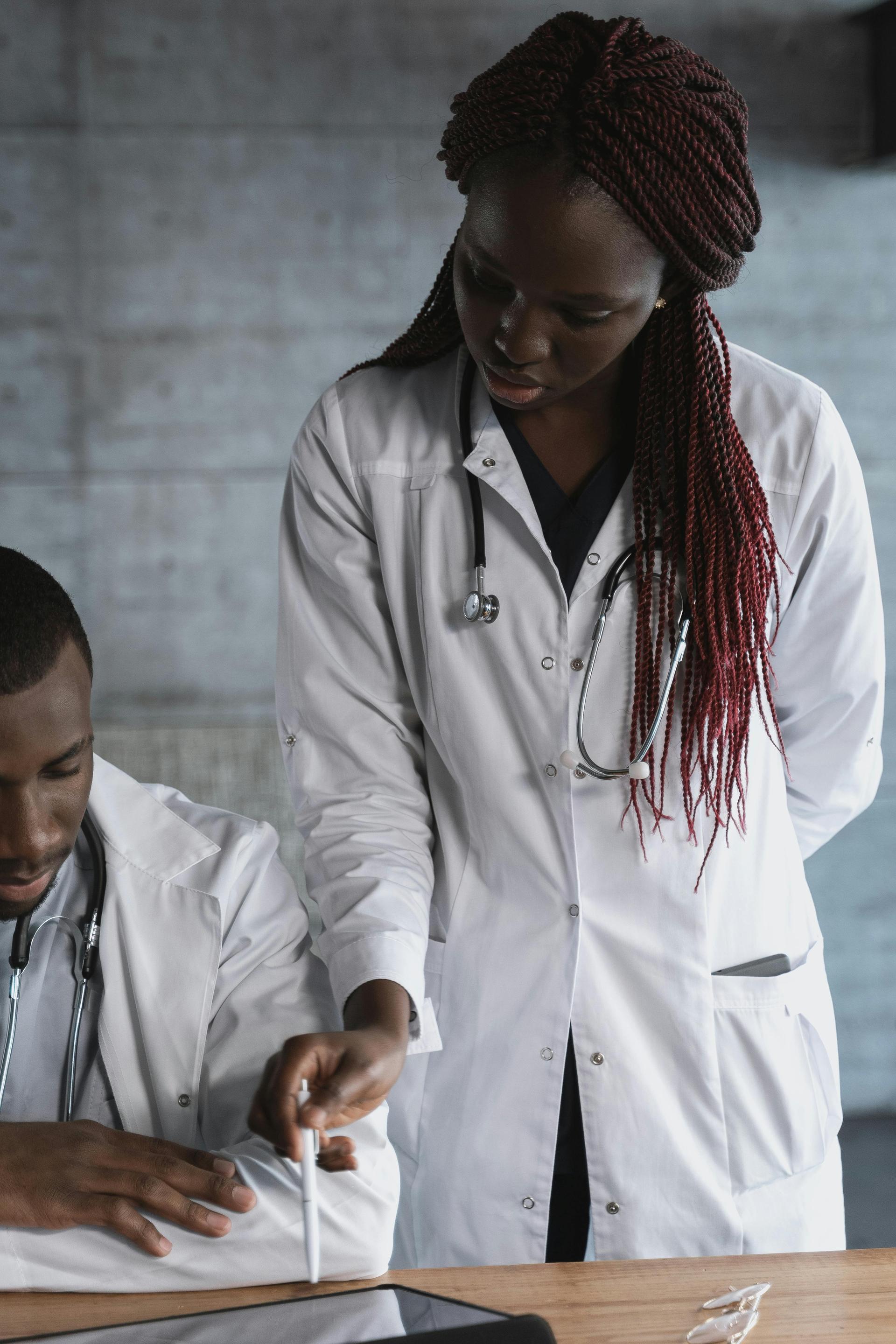 Two Black medical professionals in white coats reviewing notes together inside a hospital clinic