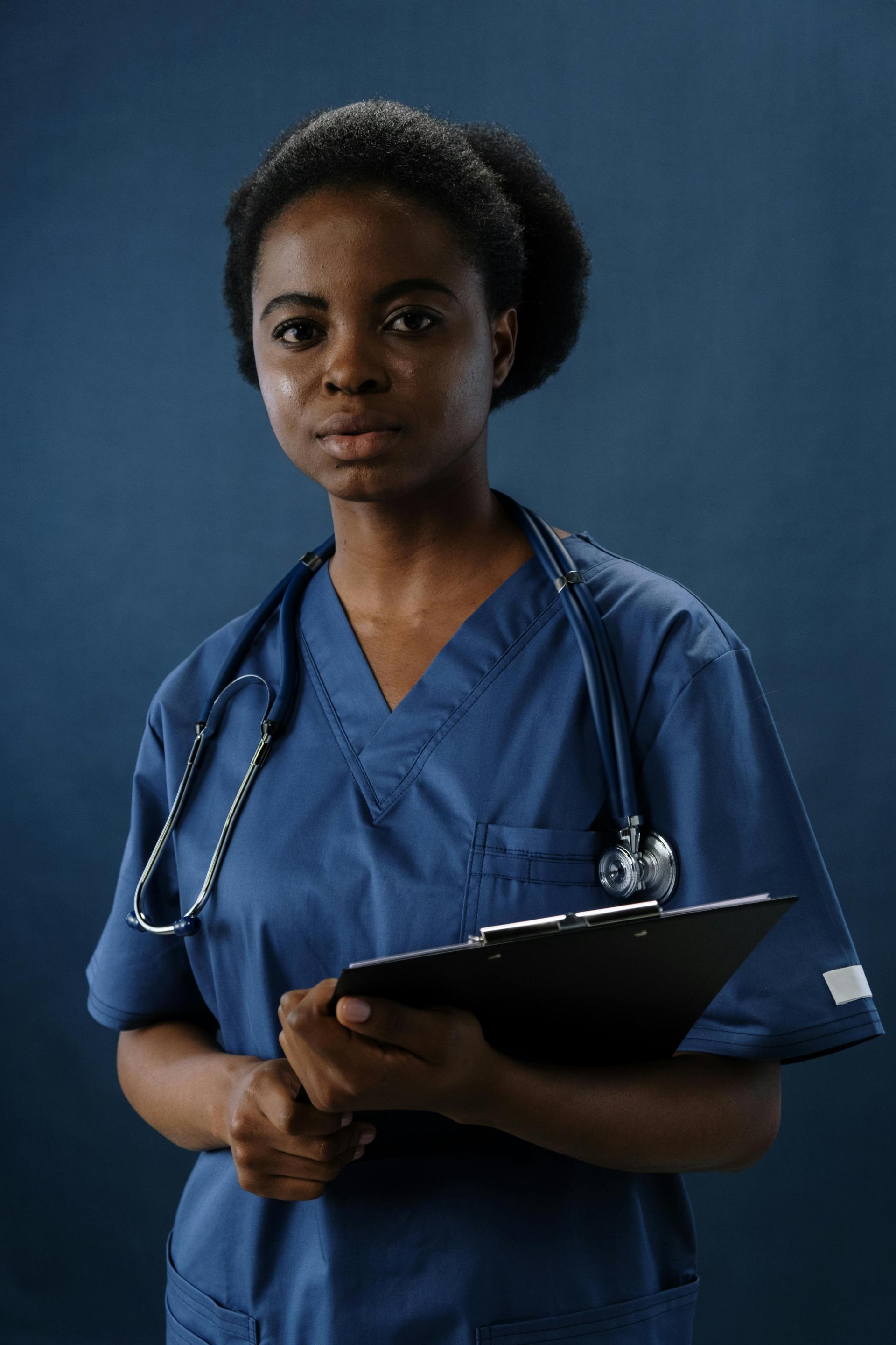 A Black nurse in blue scrubs holds a clipboard on a hospital ward, an image used to illustrate the workplace experiences of African medical workers in the United States.