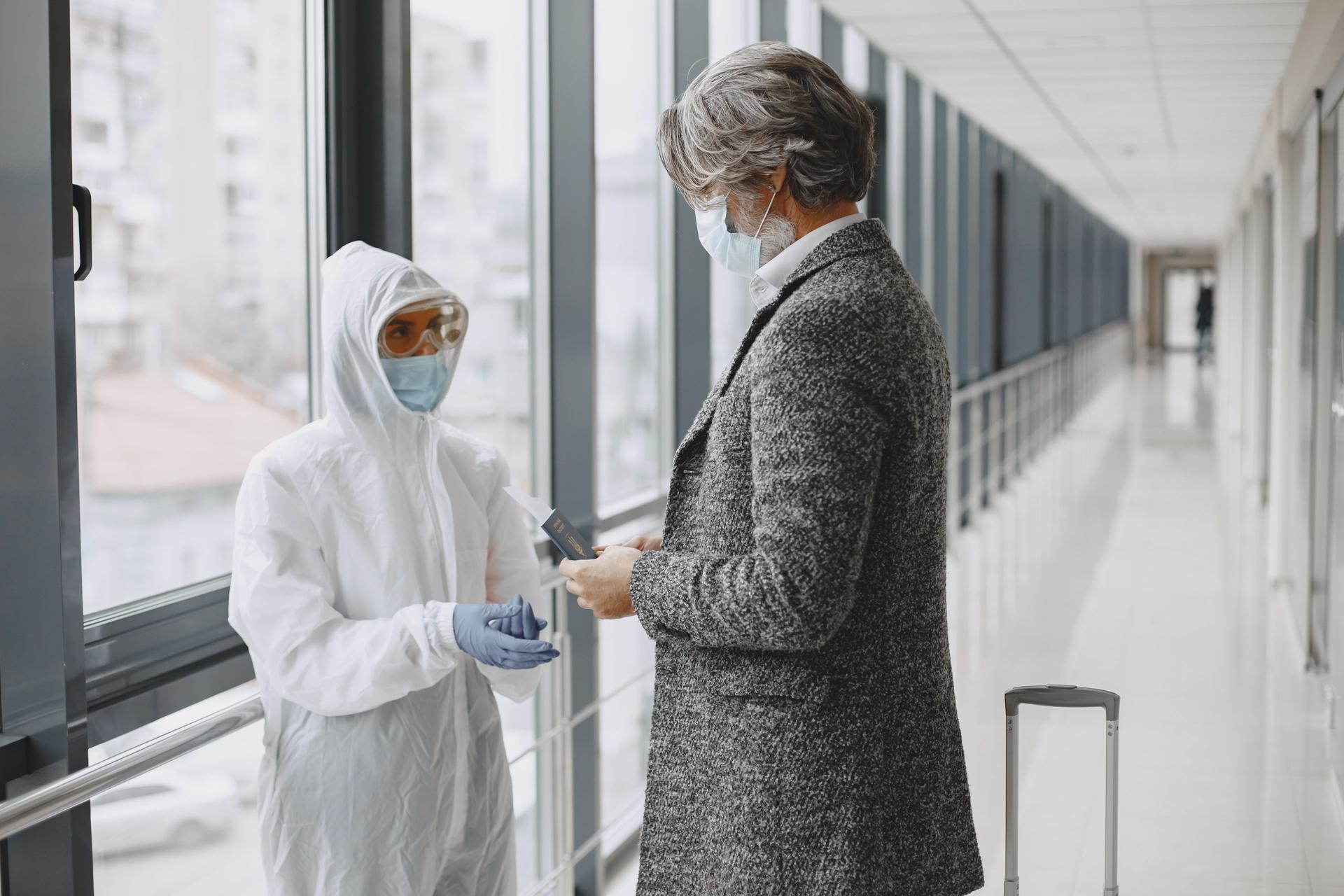 A traveler in a gray suit speaks with a masked health worker in white PPE at an airport screening station.