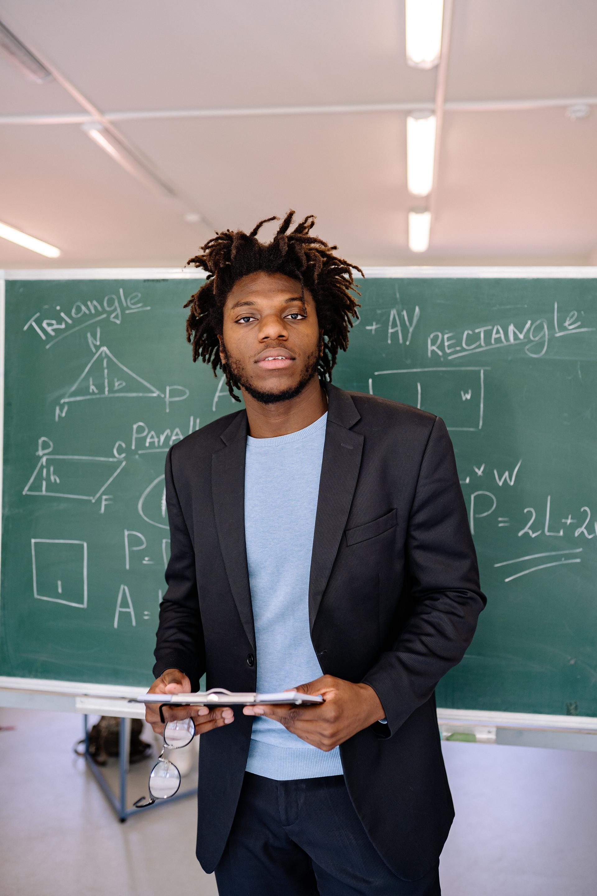 A teacher stands beside a chalkboard at the front of a classroom, marker in hand, mid-lesson.