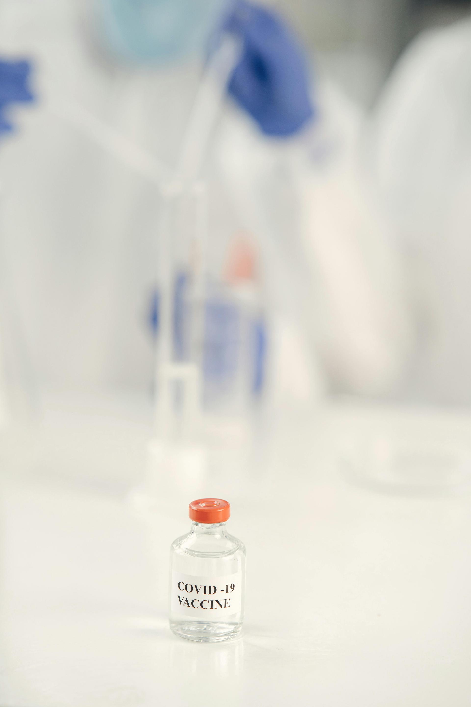 A gloved hand holds a glass vaccine ampule in a laboratory while research scientists work in the background