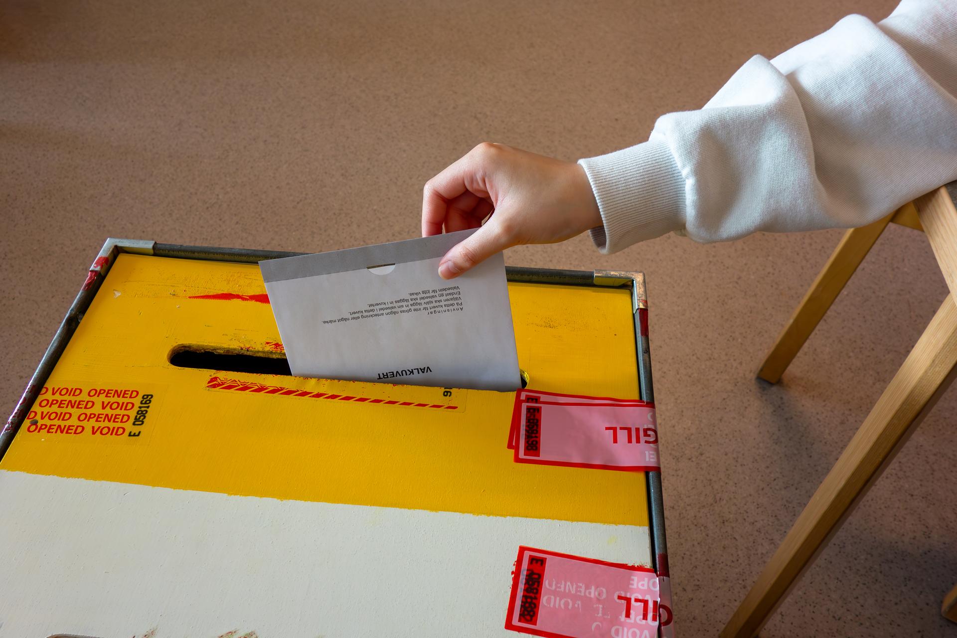 A hand drops a paper ballot into a sealed ballot box at a polling station.