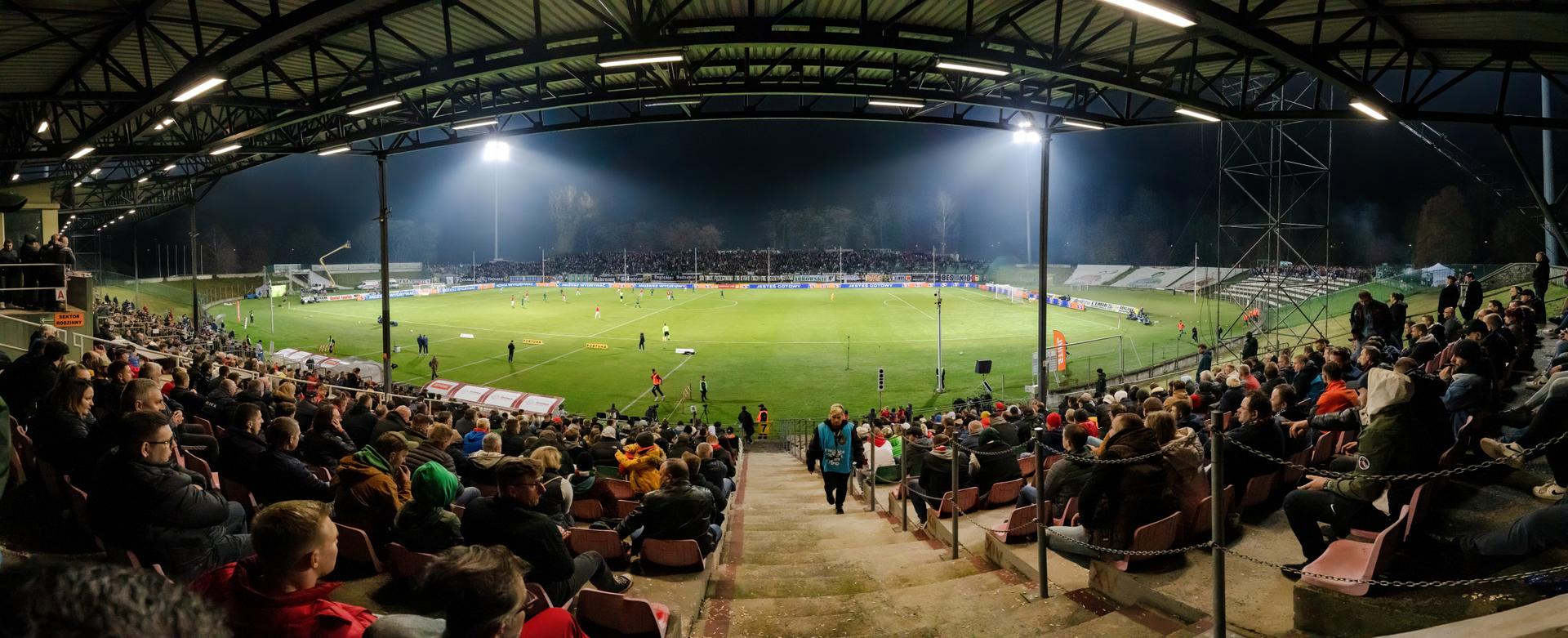 A packed football stadium crowd waving scarves and flags under floodlights during an evening match