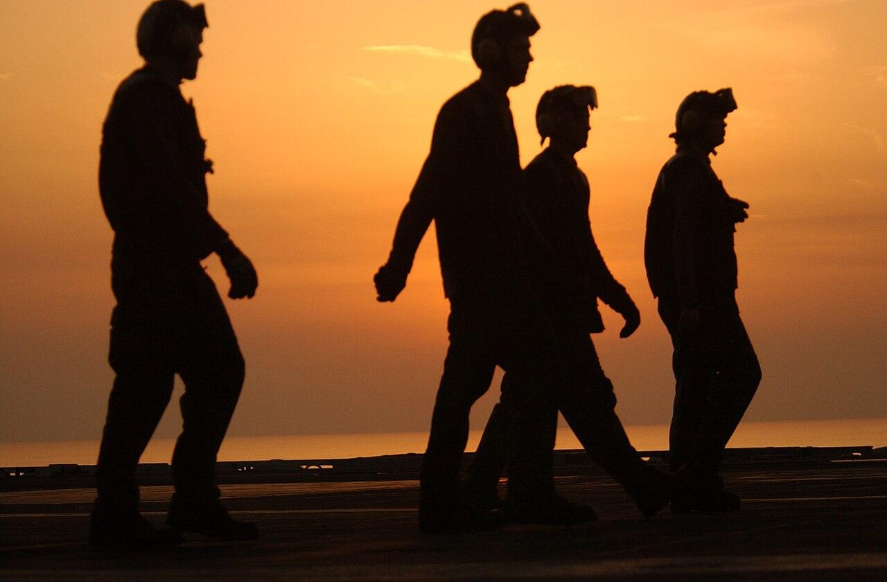 US Navy crewmembers walk across a ship's flight deck at sunset over the Arabian Sea