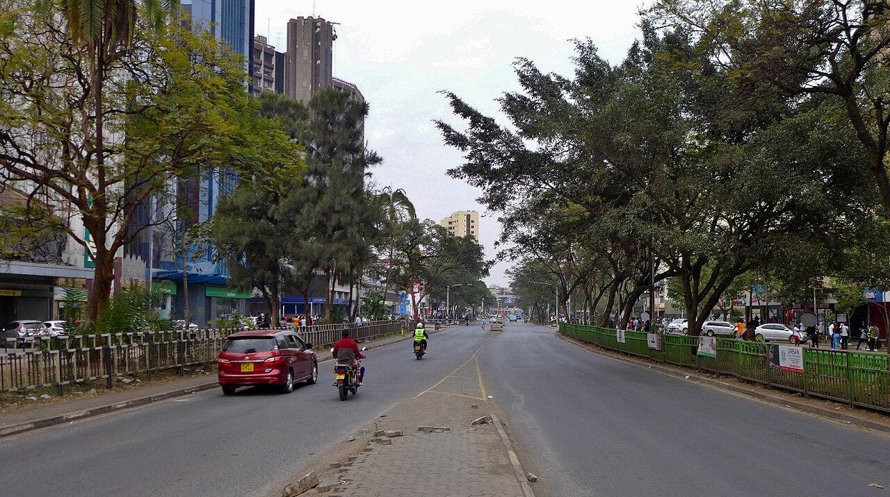A busy street scene in central Nairobi, Kenya, with pedestrians and vehicles moving past commercial buildings