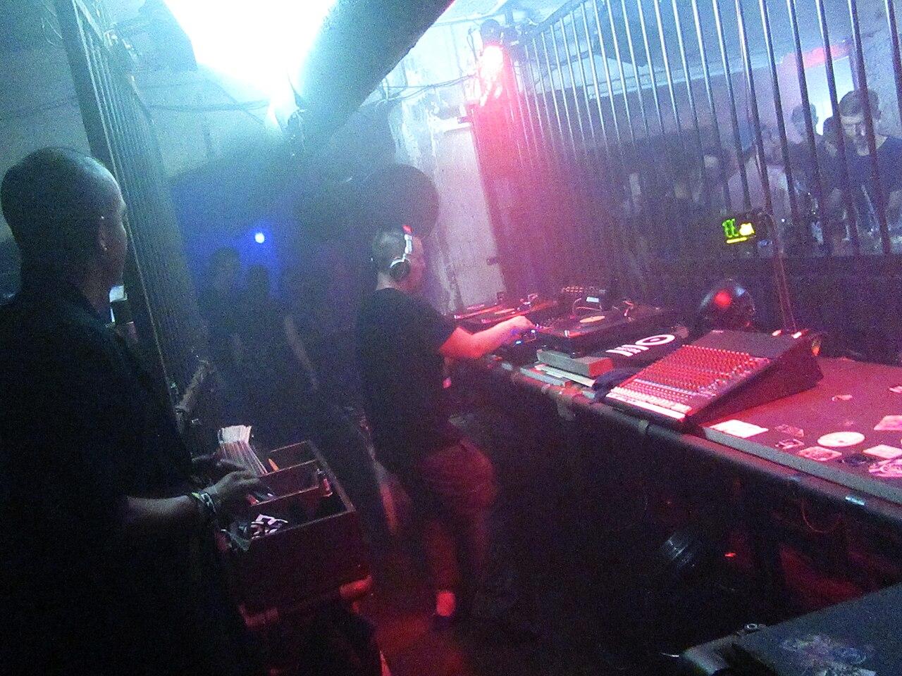 A DJ performs to a crowd inside a darkened European nightclub, lit by stage lights and a glowing booth