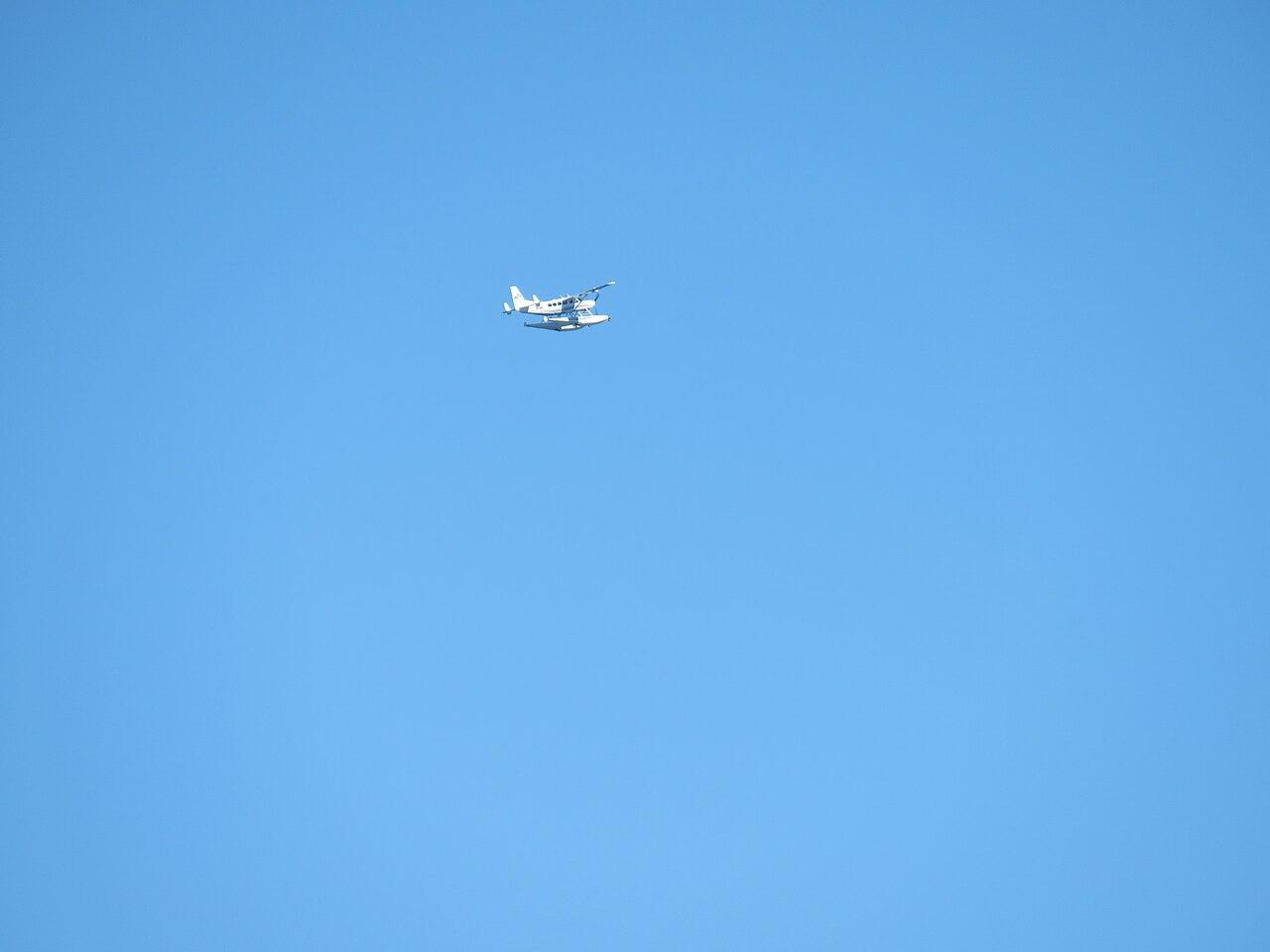 A single-engine Cessna 208 Caravan turboprop aircraft in flight over coastal water under a clear sky.