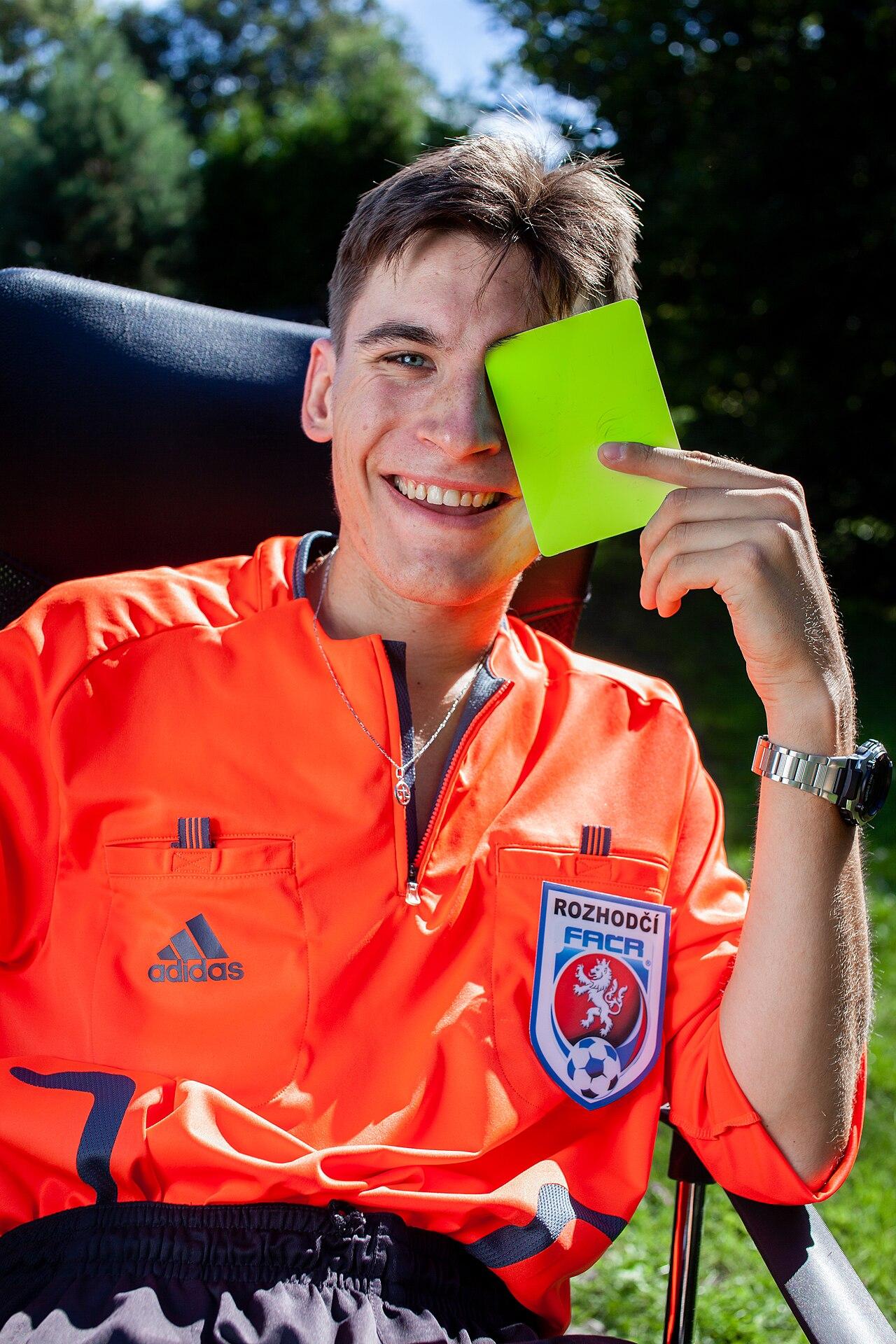 A football referee in uniform stands on the pitch holding his whistle during a match