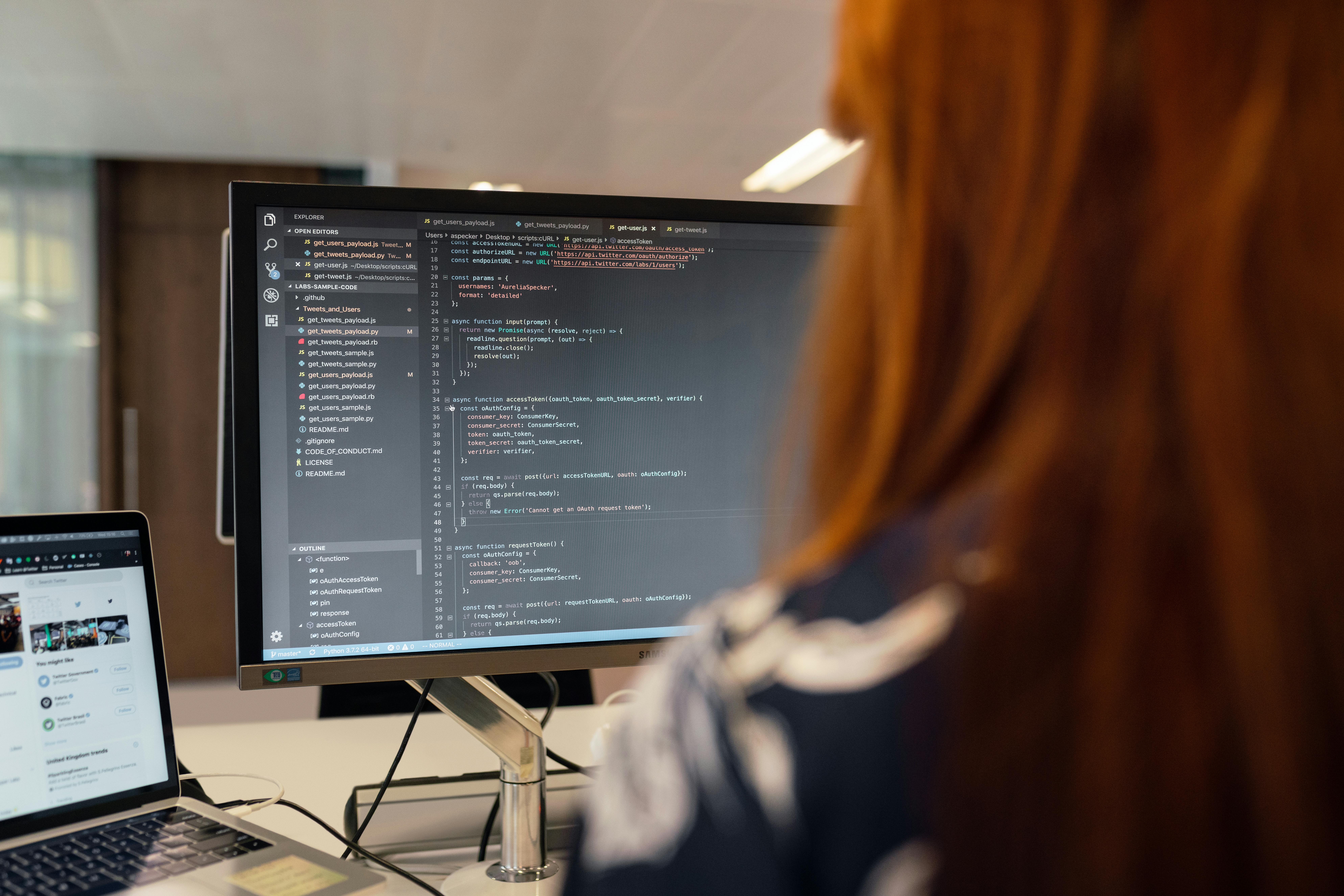 A software engineer working at a multi-monitor computer setup in a tech office, viewed from over the shoulder