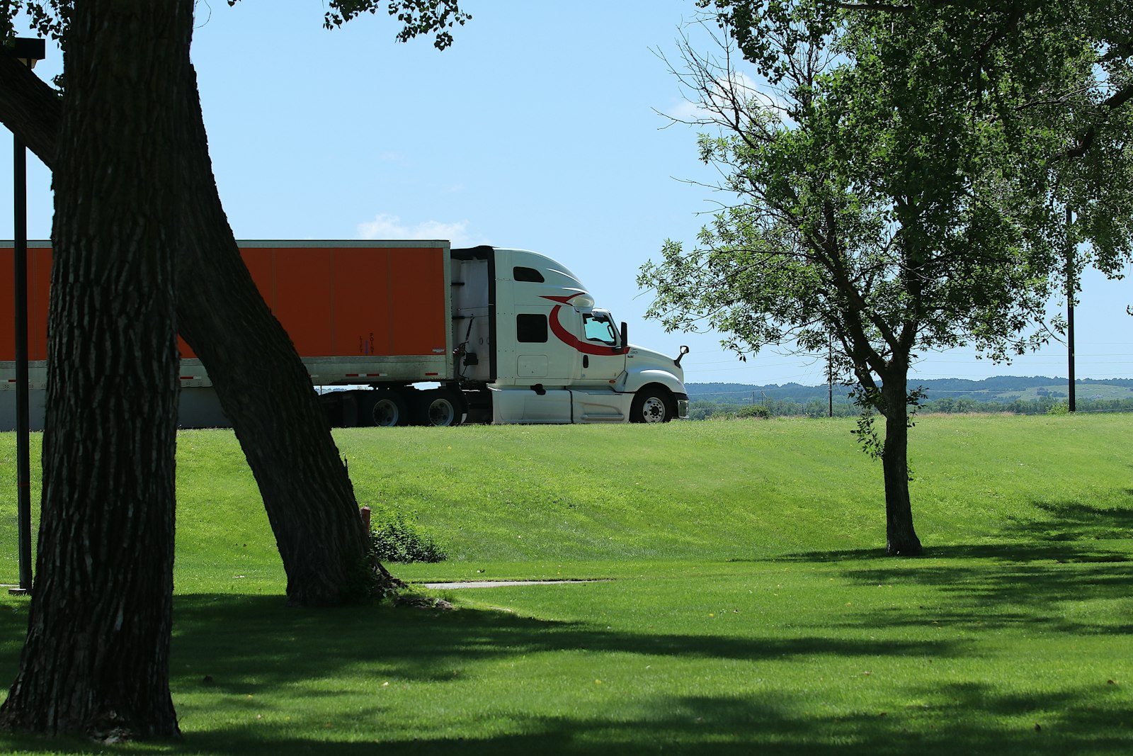 A white semi-truck with an orange trailer travels along an open American highway, illustrating the trucking industry reshaped by new federal licensing rules.