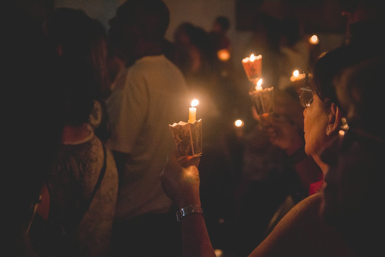 A small group holds lit candles in the dark during a vigil, hands cupped around the flames as a symbol of remembrance.