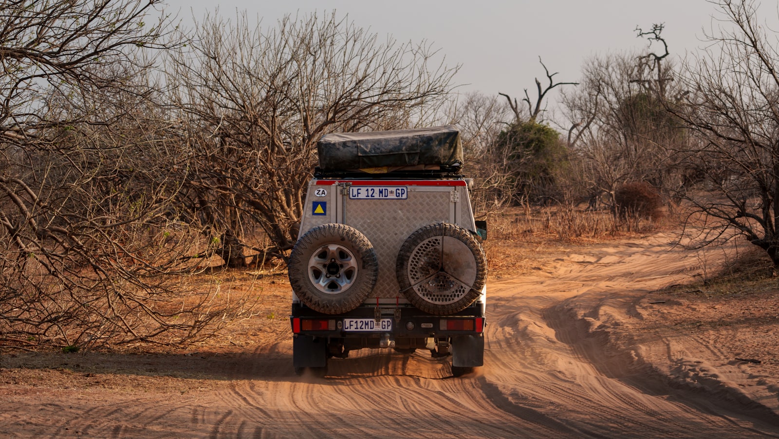 A dust-covered off-road vehicle drives along a remote dirt track, evoking a long overland journey across rough terrain.